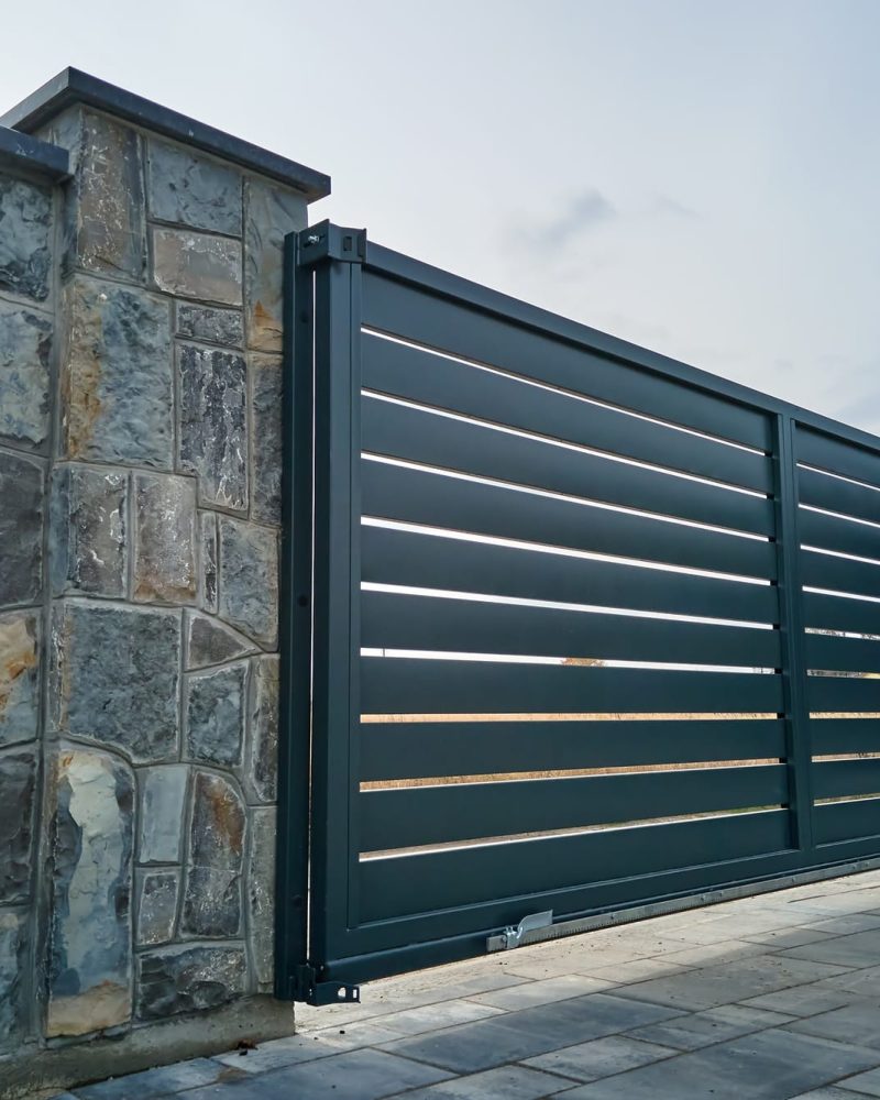 Wide automatic sliding gate with remote control installed in high stone fence wall.